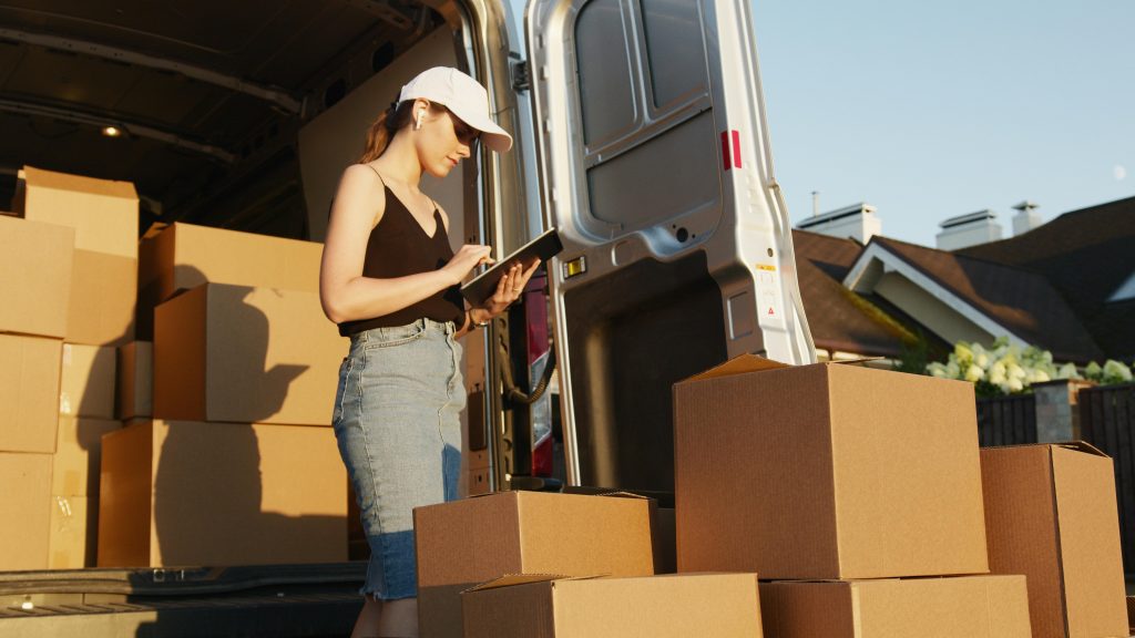 A woman using a tablet to manage deliveries with stacked cardboard boxes by a van.