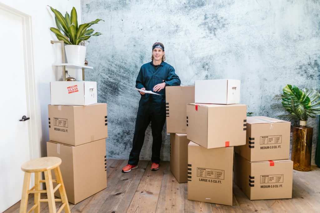 Smiling mover in coveralls standing with parcels, clipboard in hand, indoors.