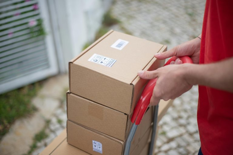 Close-up of hands holding a trolley with stacked cardboard boxes outdoors.